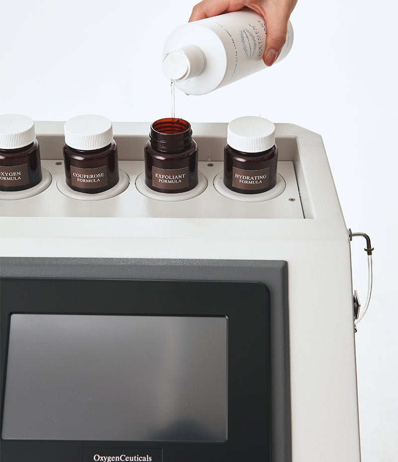 Hand pouring liquid from a white bottle into an amber jar labelled Exfoliant Formula on top of an OxygenCeuticals device with a screen